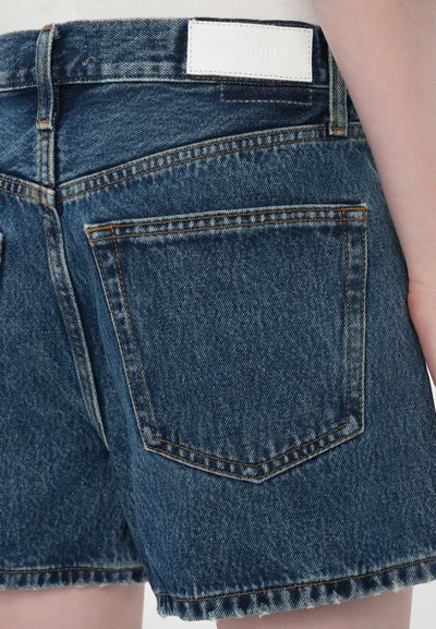 Shorts en jean bleu foncé avec un ourlet effiloché, présentant une étiquette en cuir blanc portant la mention "RE/DONE" et deux poches arrière avec des coutures contrastantes.