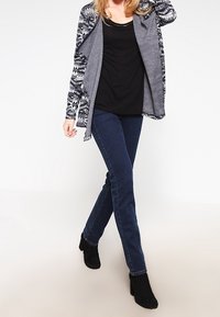 Šedý cardigan s černobílými vzorovanými rukávy, nošený přes černé tričko, spárovaný s tmavě modrými džíny a černými kotníkovými botami.