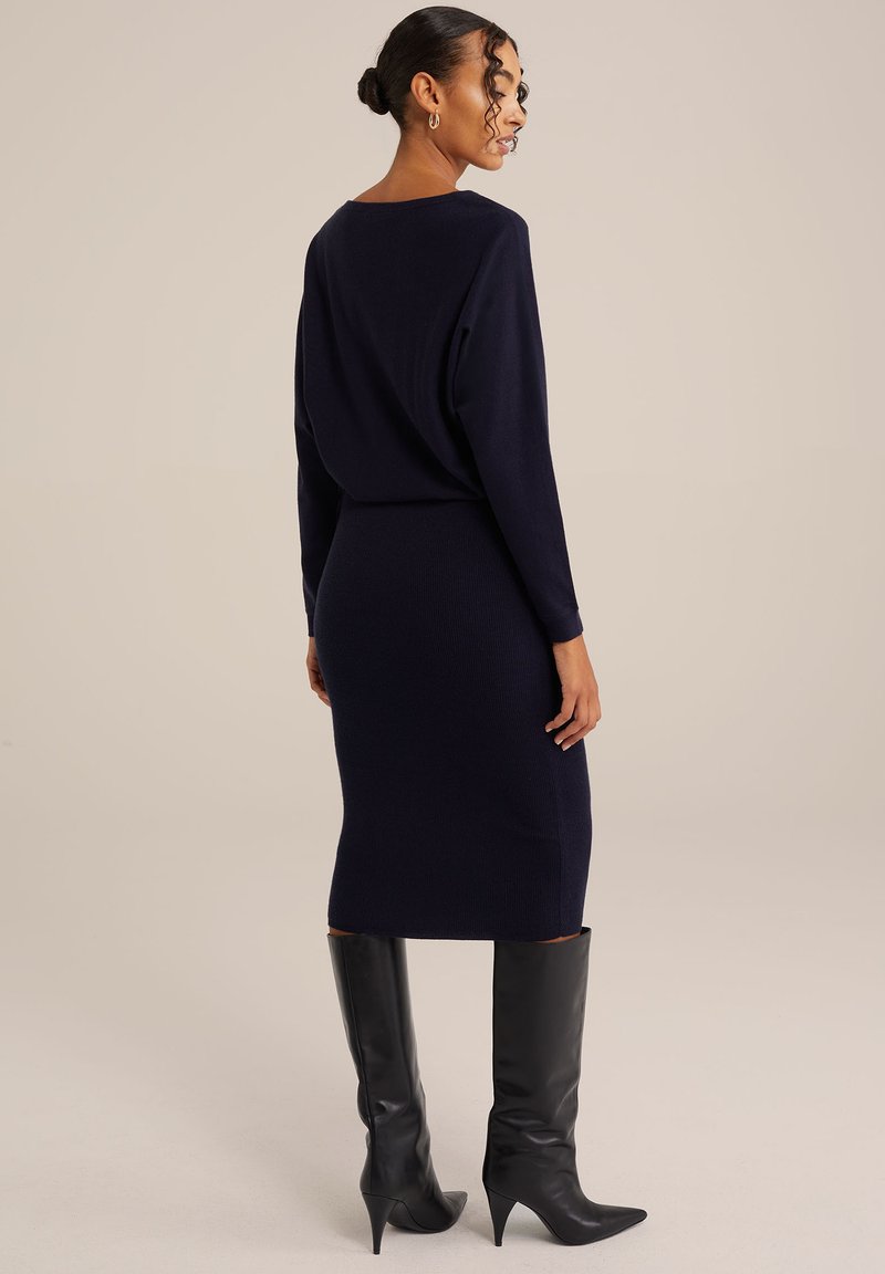 Navy-blaues Strickkleid mit langen Ärmeln, figurnaher Silhouette, knielanger Länge und rundem Ausschnitt. Dazu werden kniehohe schwarze Stiefel mit Absatz getragen.
