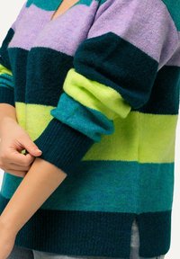 Osoba podwijająca rękaw grubej, kolorowej swetra w paski w odcieniach fioletu, ciemnej zieleni, limonki i zieleni morskiej.