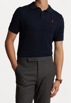 Mann trägt dunkelblaues, kurzärmeliges Polohemd mit Kabelstrickmuster und rotem Logo sowie graue, maßgeschneiderte Hose, Hand in der Tasche, braune Lederuhr am Handgelenk.