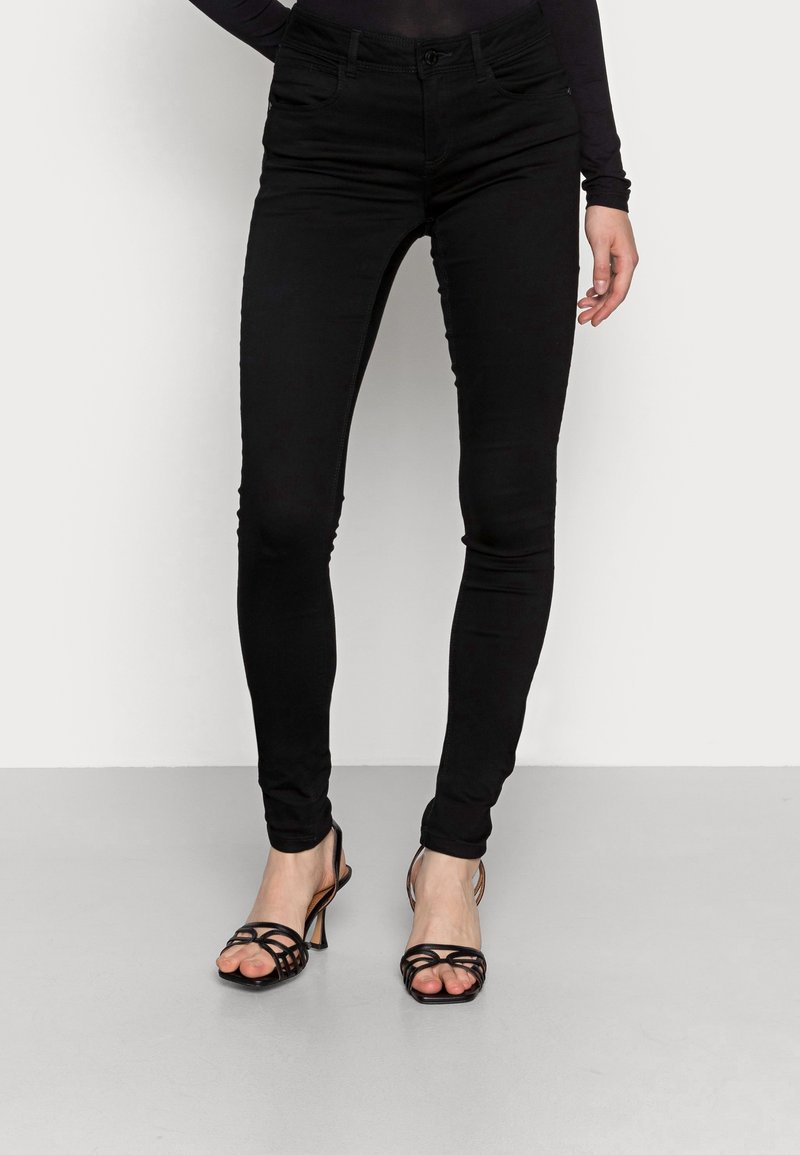 Femme portant un jean skinny noir et des sandales à talons hauts noires à lanières, debout contre un mur blanc et un sol gris.