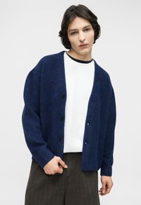 Νεαρό άτομο με μεσαίου μήκους σκούρα μαλλιά φοράει μπλε ζακέτα με υφή πάνω από λευκό πουκάμισο και σκούρο παντελόνι με φαρδιά μπότα.