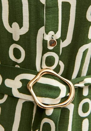 Tejido verde con un patrón de cadenas blancas, que presenta una hebilla ovalada de tono dorado y un detalle de botón redondo. Superficie texturizada en toda su extensión.