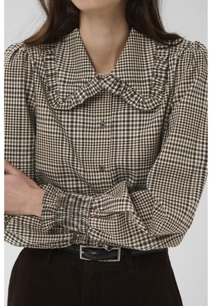 Femme portant un chemisier beige et noir à carreaux avec un grand col volanté et des manches bouffantes, associé à un pantalon foncé et une ceinture noire.
