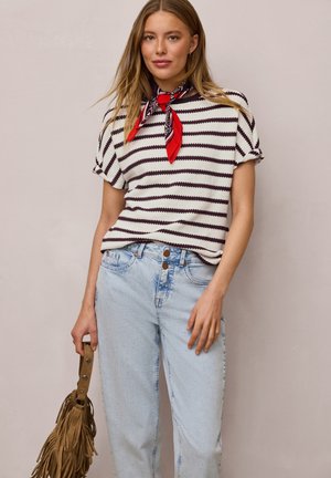 Vrouw draagt een zwart-wit gestreepte top met korte mouwen, rode patroon neksjaal, lichtblauwe hoog getailleerde jeans, houdt een bruine franjetas vast.