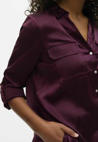 Chemise boutonnée bordeaux avec col, deux poches avant et manches retroussées, en tissu lisse avec un léger éclat.