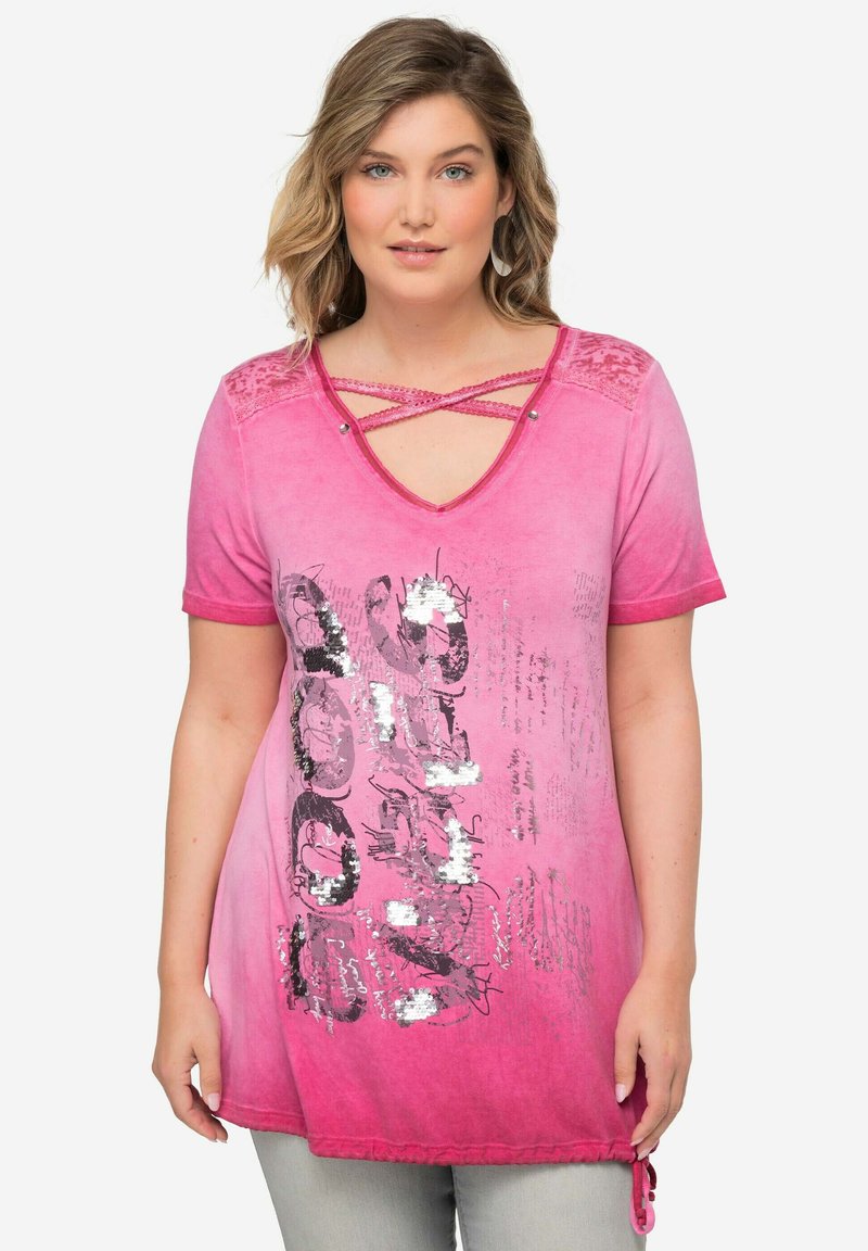 Vaaleanpunainen t-paita, jossa on syvä V-pääntie ristiin koristeluilla, paljetilla painettu graafinen kuvio ja lyhyet hihat, joissa on solmittu helma. Pehmeä kangas.