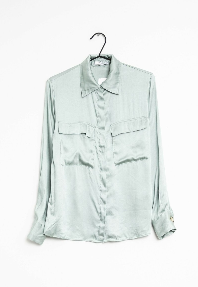 Chemise longue à manches longues en satin vert clair avec deux poches poitrine, accrochée à un cintre noir sur fond blanc.