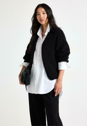 Donna con capelli lunghi e scuri indossa una camicia bianca con bottoni sotto un cardigan nero, tiene una pochette nera, in piedi davanti a uno sfondo chiaro e uniforme.