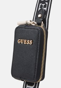 Étui zippé noir texturé avec logo "GUESS" doré, ferrures argentées, et une bandoulière rayée réglable.