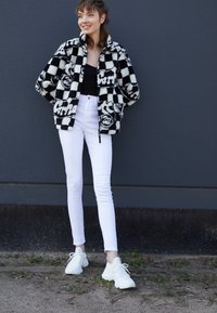 Musta-valkoinen ruudullinen fleecetakki graafisilla yksityiskohdilla, yhdistettynä valkoisiin slim-fit-farkkuihin ja valkoisiin tennareihin teksturoidulla pinnalla.