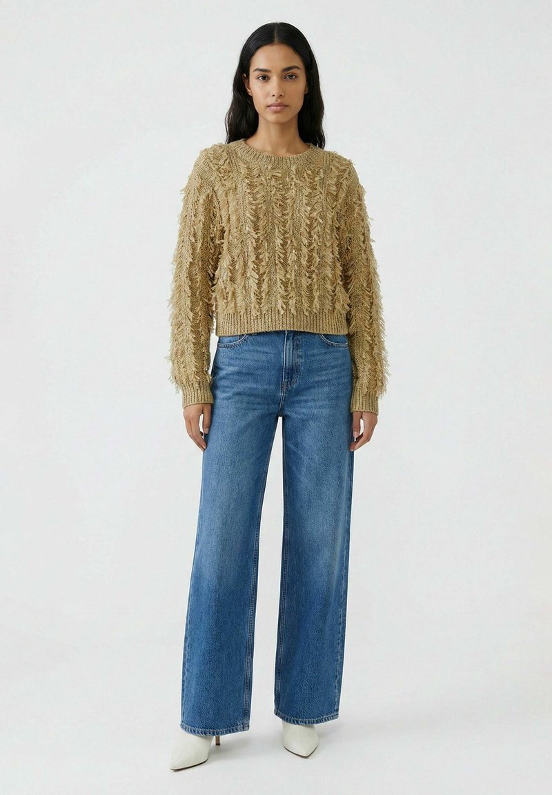 Femme portant un pull en maille texturée beige, un jean bleu à jambes larges, et des bottines blanches à bout pointu, debout devant un fond clair uni.