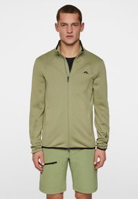 Zielonooliwkowa kurtka rozpinana z wysokim kołnierzem, dopasowanym fasonem i czarnymi akcentami. Noszona na czarnej koszulce i zestawiona z pasującymi szortami.