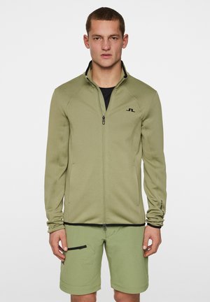 Giacca zip-up verde oliva con colletto alto, design aderente e dettagli neri. Indossata sopra una maglietta nera e abbinata a pantaloni corti coordinati.
