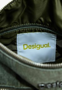 Intérieur en tissu vert et lisse avec une étiquette blanche visible affichant "Desigual" en texte jaune ; doté d'une fermeture éclair argentée.