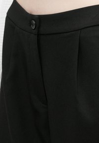 Nærbilde av svarte skreddersydde bukser med knappekant og sidelomme, på en person iført en lysfarget topp.