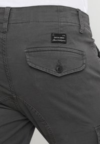 Pantalones de color gris oscuro con un bolsillo trasero que tiene un botón. Una etiqueta está cosida por encima del bolsillo, mostrando el nombre de la marca y el logo.