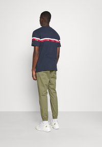 Man med marinblå t-shirt med vita och röda ränder, olivgröna joggerbyxor, vita strumpor och vita sneakers, vänd bort från kameran mot en vit bakgrund.