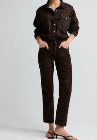 Giacca di jeans con stampa leopardata e pantaloni coordinati in marrone scuro con macchie nere. Presenta chiusure a bottoni e due tasche anteriori. Indossate sandali piatti.