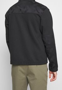 Svart jacka med mönstrade axlar och ärmar, textil med struktur, elastiska muddar och rak skärning nederst. Bärs med olivgröna byxor.