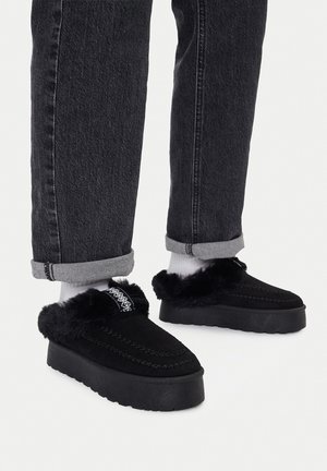 Person, die schwarze Slip-On-Plateauschuhe mit Fellfutter, weiße Socken und hochgekrempelte dunkle Jeans auf weißem Hintergrund trägt.