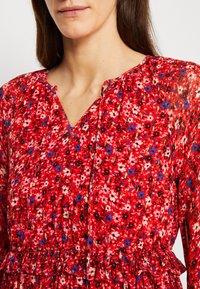 Blouse fleurie rouge avec un col en V, manches longues, détails froncés et ourlet à volants. Le tissu présente un motif de petites fleurs multicolores.