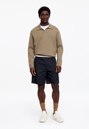 Mann steht, trägt ein beige langärmliges Polohemd, weißes Unterhemd, schwarze Shorts, beigefarbene Socken und weiße Sneakers vor weißem Hintergrund.