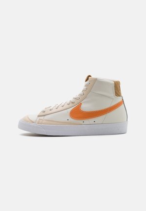 Weißer High-Top-Sneaker mit beigen Akzenten, orangefarbenem Nike-Swoosh, weißen Schnürsenkeln und weißer Sohle, seitliche Ansicht vor weißem Hintergrund.