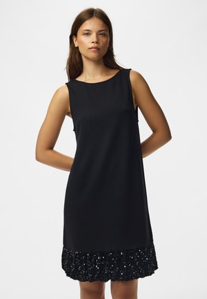 Mujer con vestido negro sin mangas con volante de lentejuelas en el dobladillo, de pie contra un fondo liso y claro.