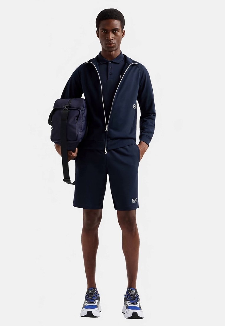 Ensemble sportif bleu marine comprenant une veste zippée et un short assorti. Comprend un polo, des baskets et un sac messager foncé.