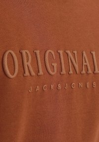 Tela de algodón naranja con letras en relieve "ORIGINAL" y "JACK&JONES". Textura suave, diseño simple, sin patrones ni acentos adicionales.