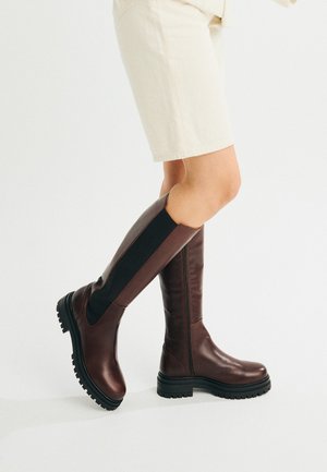 Jambes chaussées de bottes en cuir marron foncé montantes jusqu'aux genoux avec des panneaux élastiques noirs et des semelles noires épaisses, assorties à un short beige clair.