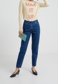 Pull beige avec "MAX IT UP" en rose, jean en denim bleu taille haute, et escarpins en cuir verni noir. Tenant un portefeuille vert menthe.