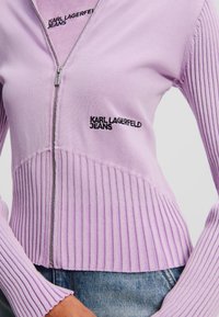 Lila sveter na zips s rebrovanou textúrou, so vyšitou logom "Karl Lagerfeld Jeans". Zips na prednej strane a priliehavé rukávy.
