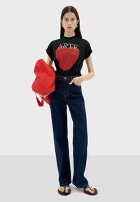 Zwarte t-shirt met rode hart en "ARTE" tekst, high-waisted blauwe denim jeans, rode tas en bruine bontsandalen. Model staat tegen een neutrale achtergrond.