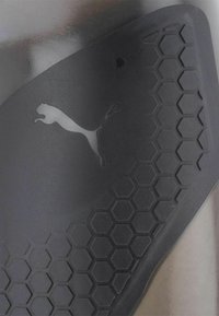 Sort beskyttelsesudstyr med et hexagonalt mønster, der har et subtilt Puma-logo. Glat overflade med mat finish og buet design.