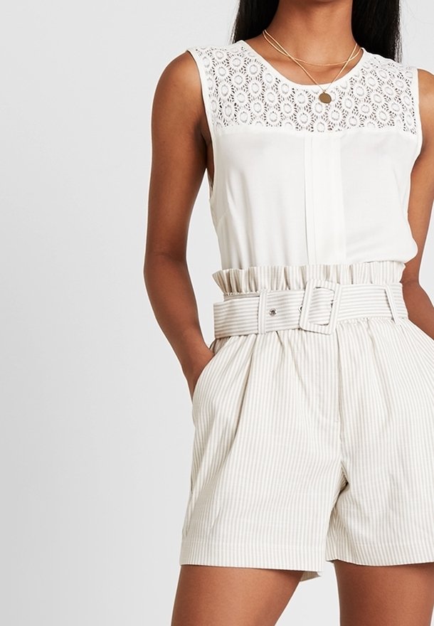 Femme portant un haut blanc sans manches avec un détail en dentelle et un short rayé taille haute avec une large ceinture, debout les mains dans les poches.