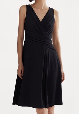 Femme portant une robe noire sans manches avec un col en V et des détails plissés autour de la taille et de la jupe.