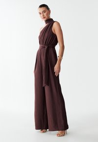 Jumpsuit sans manches bordeaux avec un col haut et une taille nouée. Présente un tissu texturé et une coupe ample. Des sandales métalliques complètent le look.