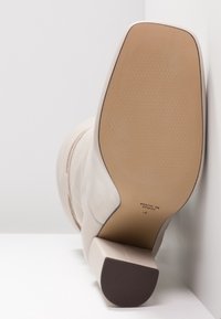 Bottine en cuir beige avec un talon carré, une texture lisse et des coutures minimales. La semelle présente un léger motif antidérapant.