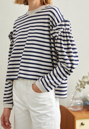 Personne portant un haut à manches longues rayé bleu marine et blanc avec des boutons en perle aux manches et un pantalon blanc, debout à l'intérieur.
