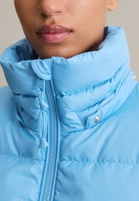Hellblaue Steppjacke mit hohem Kragen, Reißverschlussdetails und verstellbarem Kordelzug. Sie hat eine gesteppten Textur und Druckknopf-Akzenten.