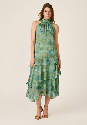 Mujer con vestido sin mangas en capas con estampado floral verde y azul con cuello halter y sandalias de tacón alto color beige, de pie frente a un fondo liso.