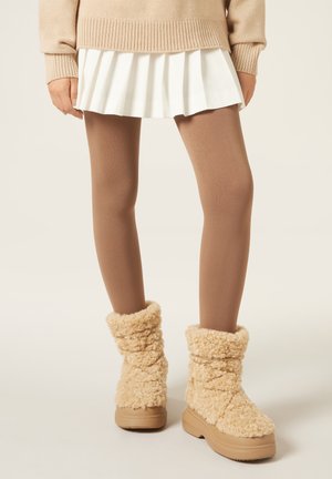 Beige flauschige Stiefeletten mit einer strukturierten Oberfläche, kombiniert mit braunen Strumpfhosen und einem plissierten weißen Minirock, dazu ein gerippter beiger Pullover.