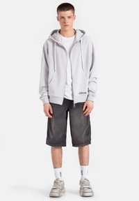 Jasnoszara bluza z kapturem na zamek błyskawiczny, z kapturem z troczkami, zestawiona z czarnymi szortami dżinsowymi i masywnymi srebrnymi sneakersami. Białe skarpetki z widocznym napisem.