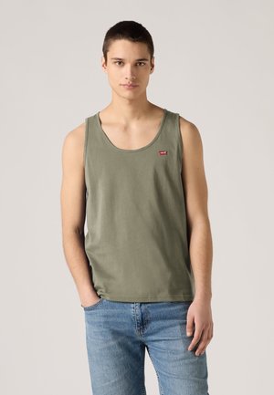 Young man with short dark hair wearing olive green tank top and light blue jeans, standing with one hand in pocket.