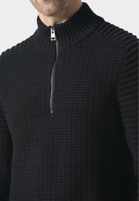 Czarny pulower z dzianiny z prążkowanym kołnierzem i zamkiem błyskawicznym. Teksturowana tkanina posiada wzorzystą strukturę na ciele i rękawach.