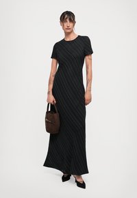 Woman with dark hair in a black, short-sleeve, floor-length striped dress holding a brown handbag and wearing black pointed shoes.