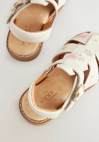 Sandale albe din piele cu broderie florală în roz și verde, tălpi căptușite și curea cu cataramă. Talpă exterioară maro cu detaliu de cusături albe.